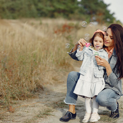 Familie in einem Herbstpark. Mutter in rosa Hemd und blauer Jeansjacke zeigt einem  fünfjährigen Mädchen, wie man Seifenblasen macht.
