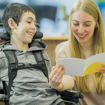 Ein Junge im Grundschulalter, mit einer körperlichen Behinderung, ist mit seiner Betreuerin in der Schule. Sie lesen zusammen ein Buch.